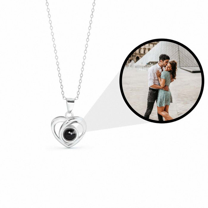 A silver heart pendant necklace with a black center stone on a white background, with a circular inset showing a couple's photo that appears inside the pendant.