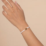 A delicate white beaded bracelet with a small gold heart-shaped charm containing a dark stone, worn on a person's wrist against a solid beige background.