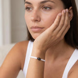 Close-up portrait of a person resting their face on one hand, wearing a white sleeveless top and a white braided bracelet with a black bead. The person has a neutral expression and is looking off to the side in soft natural light.