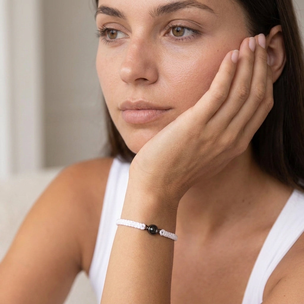 Close-up portrait of a person resting their face on one hand, wearing a white sleeveless top and a white braided bracelet with a black bead. The person has a neutral expression and is looking off to the side in soft natural light.