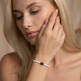 Close-up portrait of a person resting their face on one hand, wearing a white sleeveless top and a white braided bracelet with a black bead. The person has a neutral expression and is looking off to the side in soft natural light.