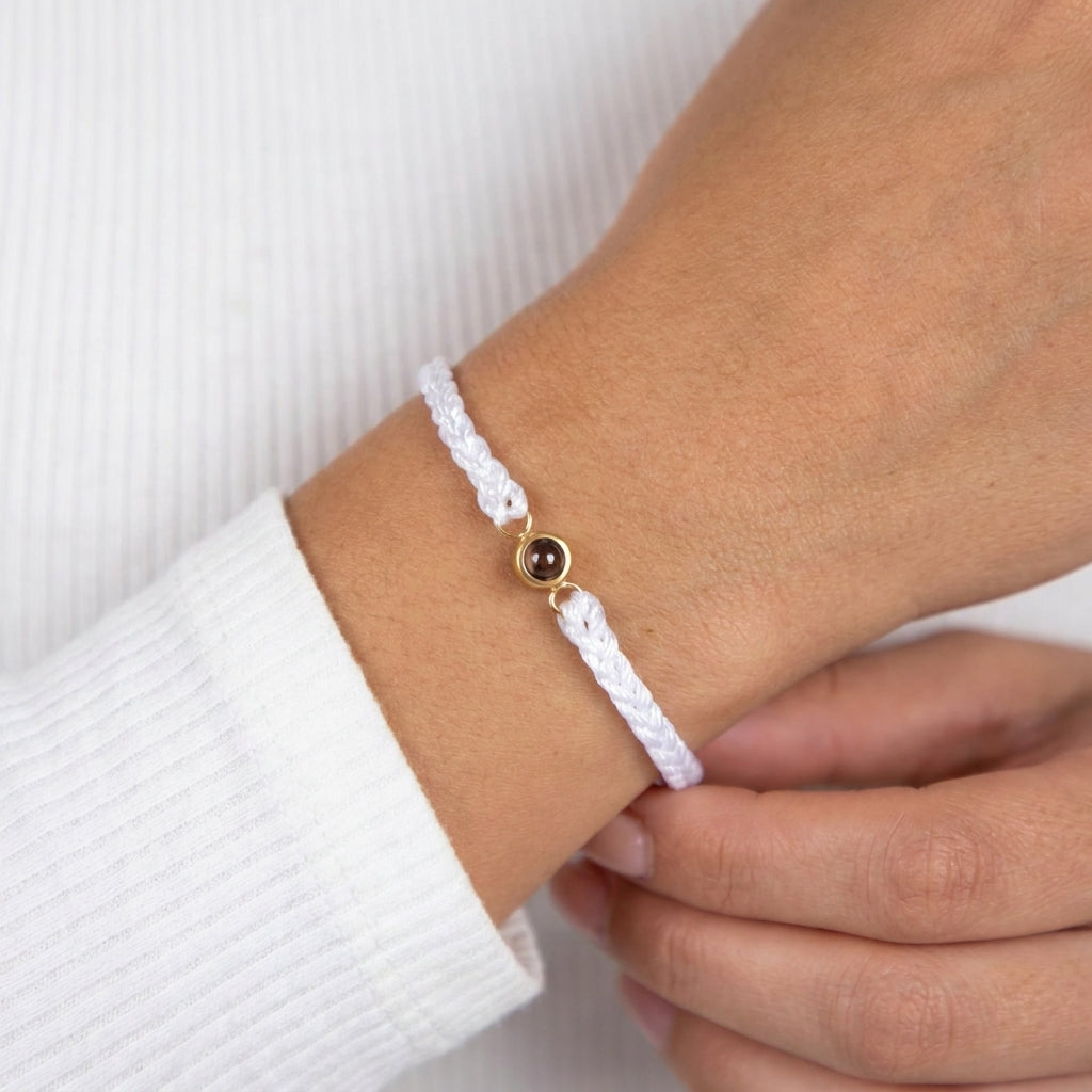 A close-up of a person's wrist wearing a delicate white braided cord bracelet with a small gold-toned circular charm containing dark brown inclusions.