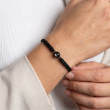 A close-up of a person's wrist wearing a black braided cord bracelet with a small round black bead centerpiece, paired with a white ribbed top and a light beige sleeve.