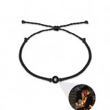 A black braided cord bracelet with a central circular charm containing a small pink stone, displayed on a white background with a circular inset photo of a couple at night.