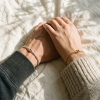 Two hands resting together on linen in sunlight, wearing matching silver and gold Love Knot Bracelets