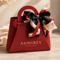A red structured gift bag with a top handle, printed with the brand name 'ADMIRES JEWELLERY' in gold, and decorated with a large black, red, and white satin bow featuring a gold charm, set against a warm neutral background.