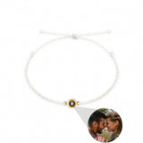 A white braided cord bracelet with a gold-toned circular charm holding a small photo of a couple, shown on a plain white background with an inset demonstrating the photo insertion.