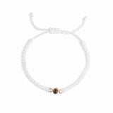 A delicate white braided cord bracelet with an adjustable sliding knot and a small rose gold circular charm featuring a dark center, shown on a plain white background.