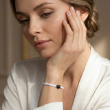 Close-up portrait of a person resting their face on one hand, wearing a white sleeveless top and a white braided bracelet with a black bead. The person has a neutral expression and is looking off to the side in soft natural light.