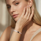 Close-up portrait of a person resting their face on one hand, wearing a white sleeveless top and a white braided bracelet with a silver bead. The person has a neutral expression and is looking off to the side in soft natural light.