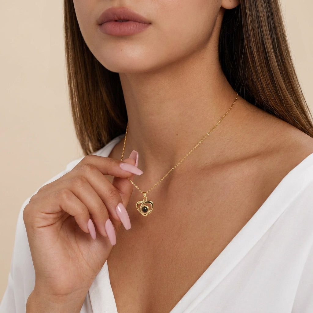 A close-up of a woman wearing a delicate gold necklace with a heart-shaped evil eye pendant, featuring a black center surrounded by small gold dots. She is wearing a white top against a soft beige background.