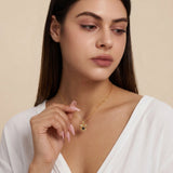 A woman wearing a delicate gold chain necklace with a small gold heart-shaped pendant featuring a black center, posed against a soft beige background.