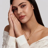 A close-up of a woman wearing a delicate gold necklace with a small heart-shaped pendant featuring a dark center, styled with a white off-the-shoulder blouse against a light gray background.