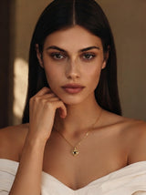 A close-up portrait of a woman wearing a delicate gold necklace with a small heart-shaped pendant featuring a dark center, styled with a white off-the-shoulder top against a warm, softly lit background.